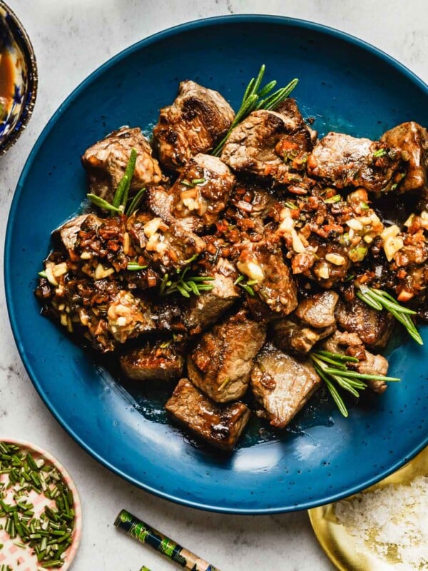 Feature image shows tender garlic steak bites top with loads of garlic and garnished with rosemary, served on a blue color serving plate.