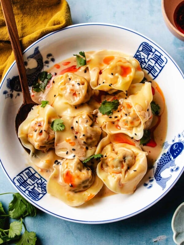 An overhead feature image shows a plate of juicy chicken wontons with spicy sesame sauce served in a blue white color bowl.