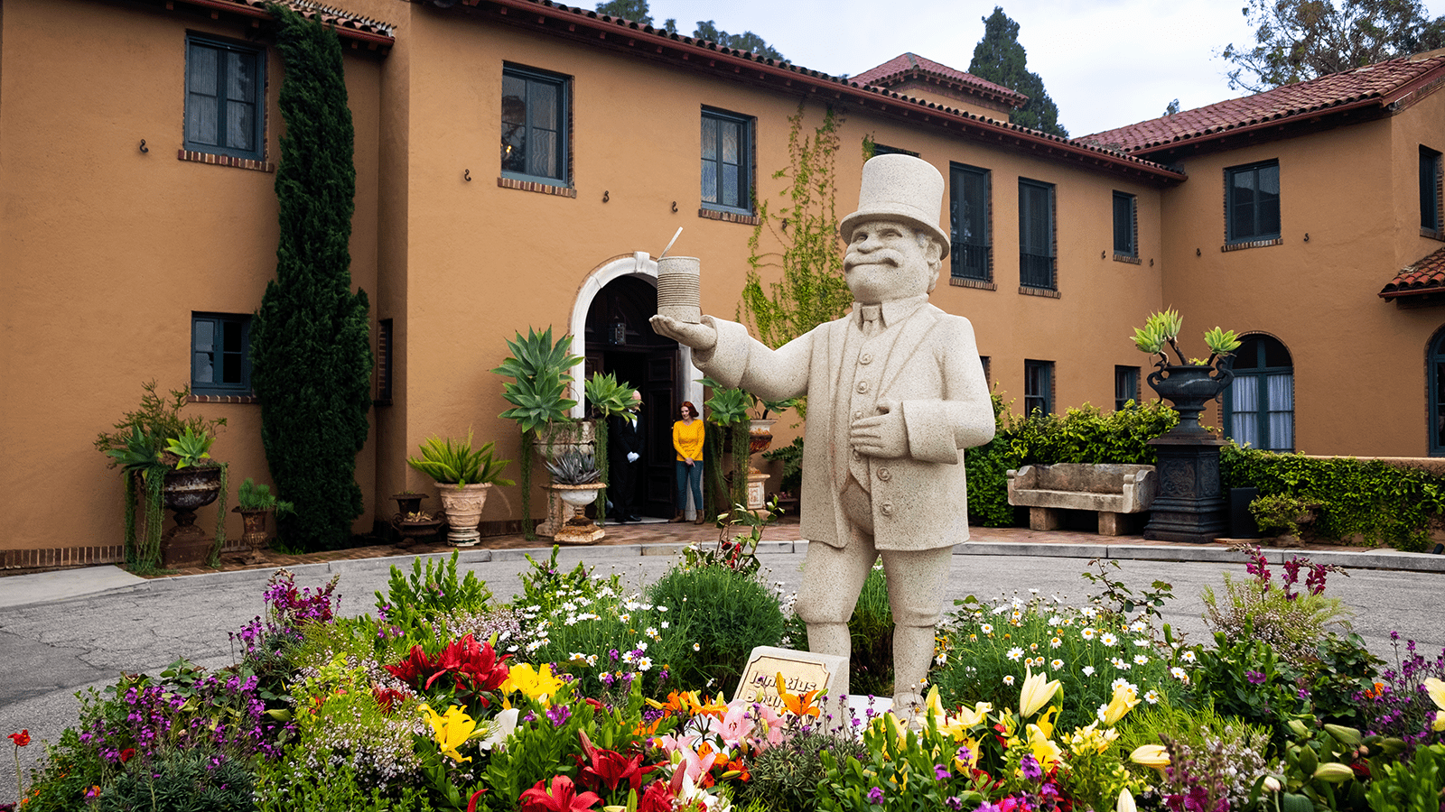 Metacore Merge Mansion Unlocked A stone statue of one of the Merge Mansion game characters is in the middle of a lush garden in front of a large mansion. The character in the garden is of an older man wearing a suit and a top hat holding an can in the air.