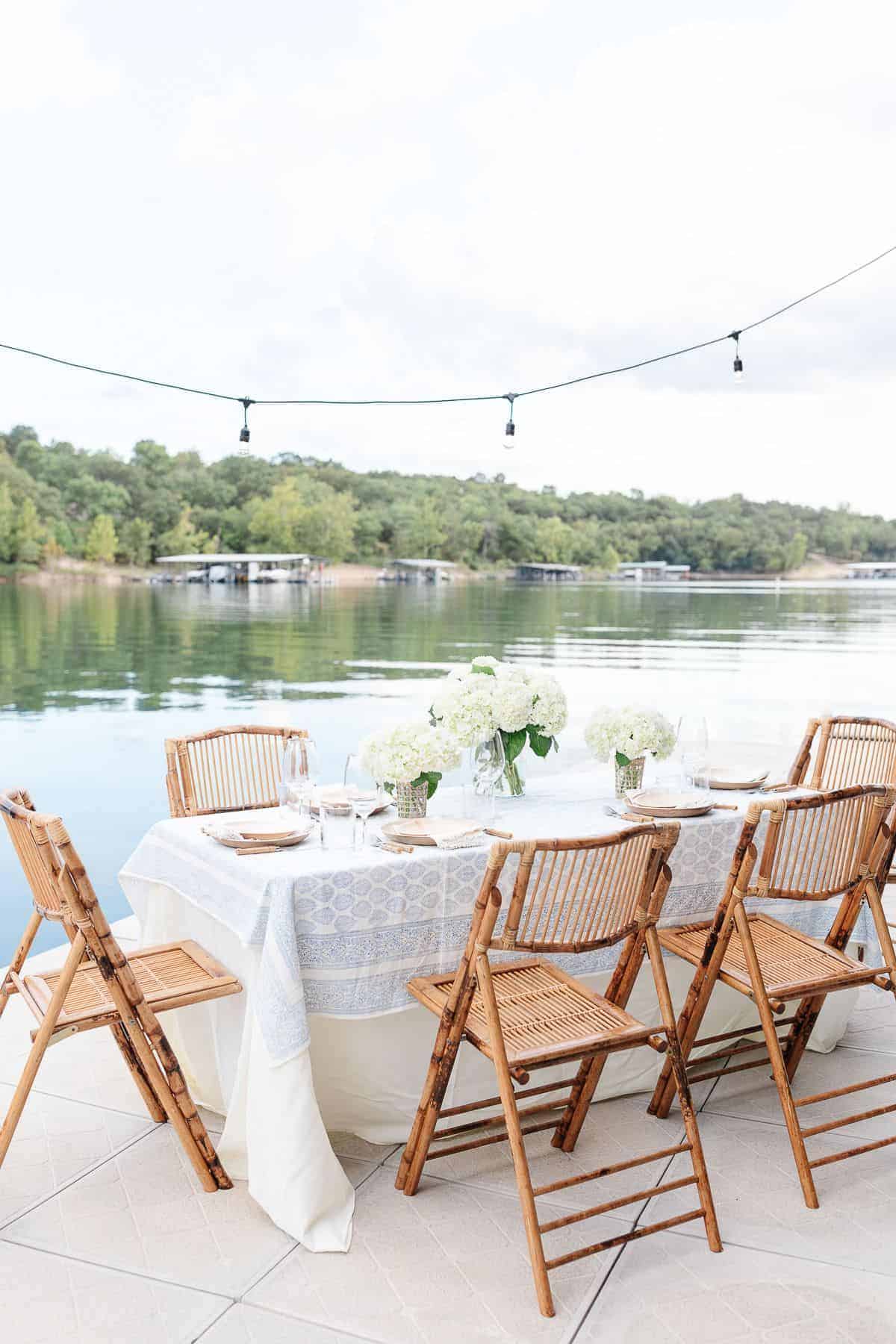 An al fresco dining table set with white flowers and a blue and white table cloth, water in the background.