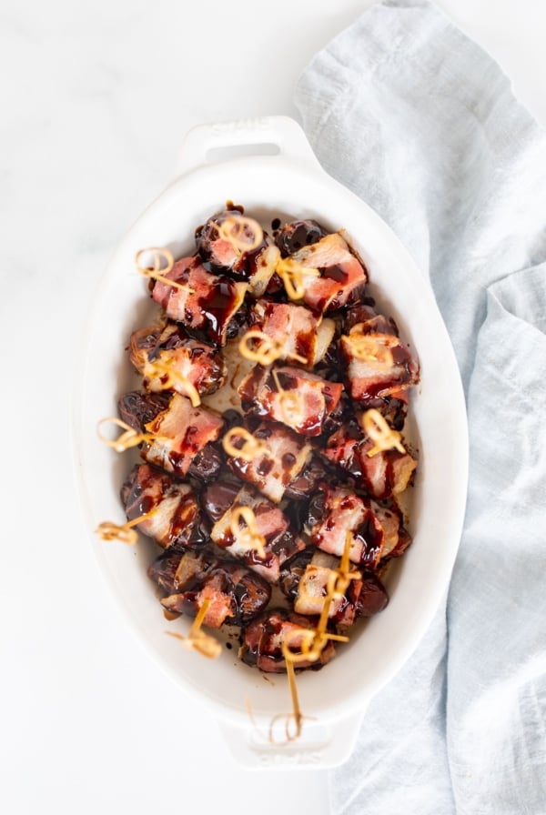 A white oval baking dish with bacon wrapped dates, each has a toothpick for serving.