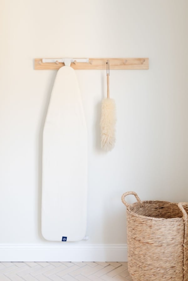 A peg rail on a white wall holding an ironing board and feather duster.