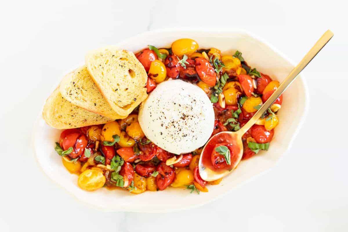 Burrata Bruschetta in a white serving bowl, crostini and a gold spoon to the side.