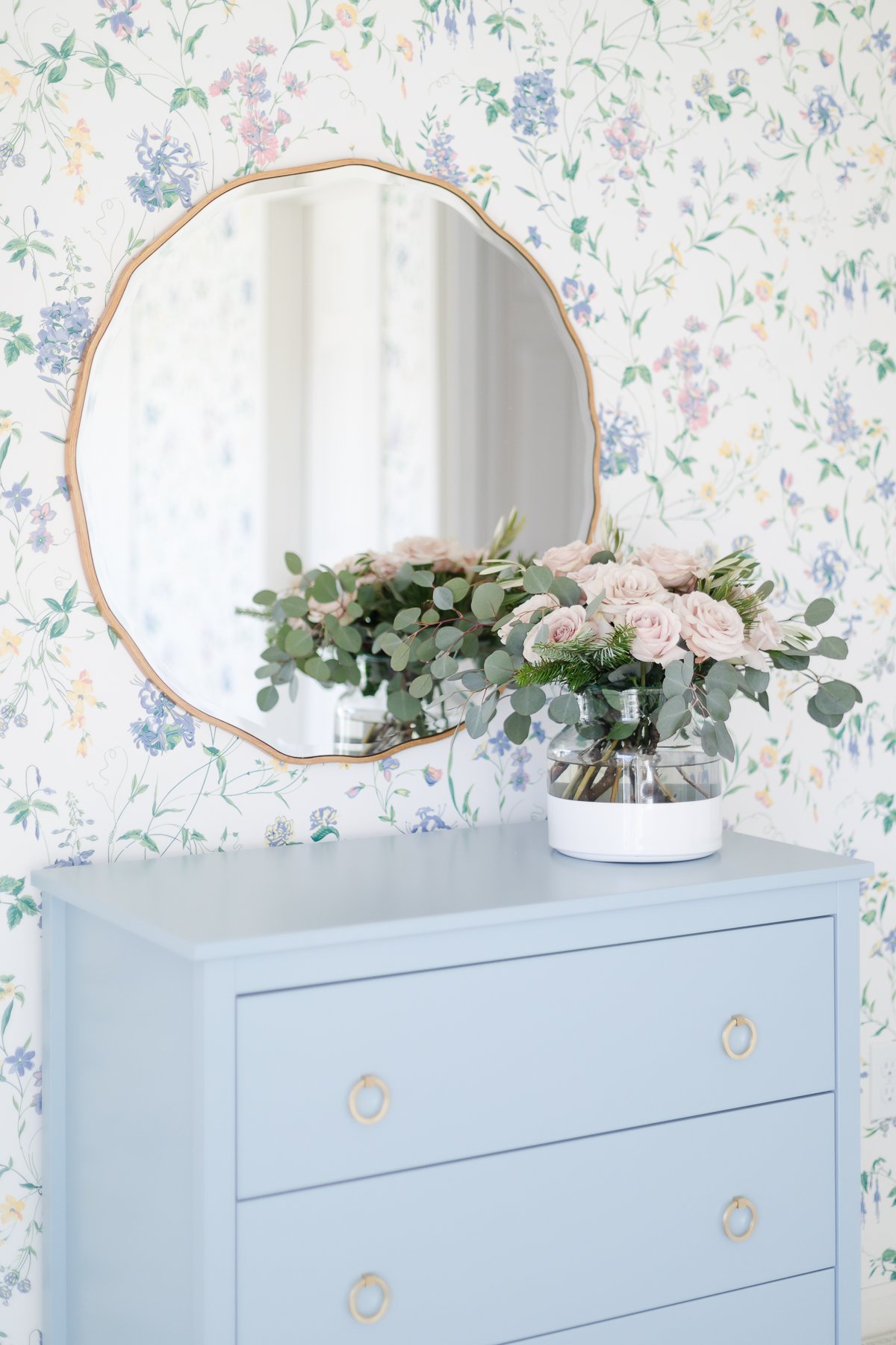 A wallpapered bedroom with a blue dresser and a gold scalloped mirror.