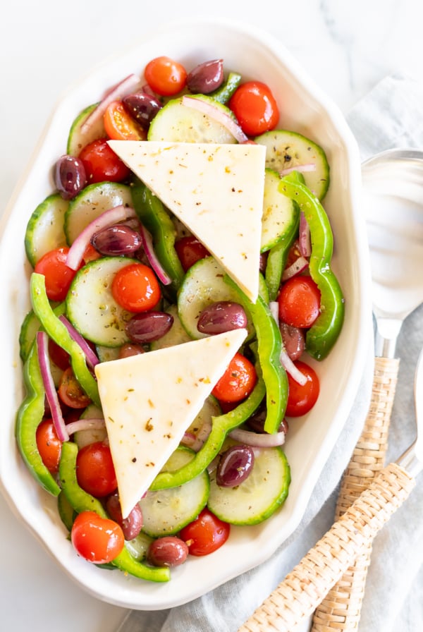 Overhead view of Greek salad on platter with two wedges of feta set over the top, next to serving spoons