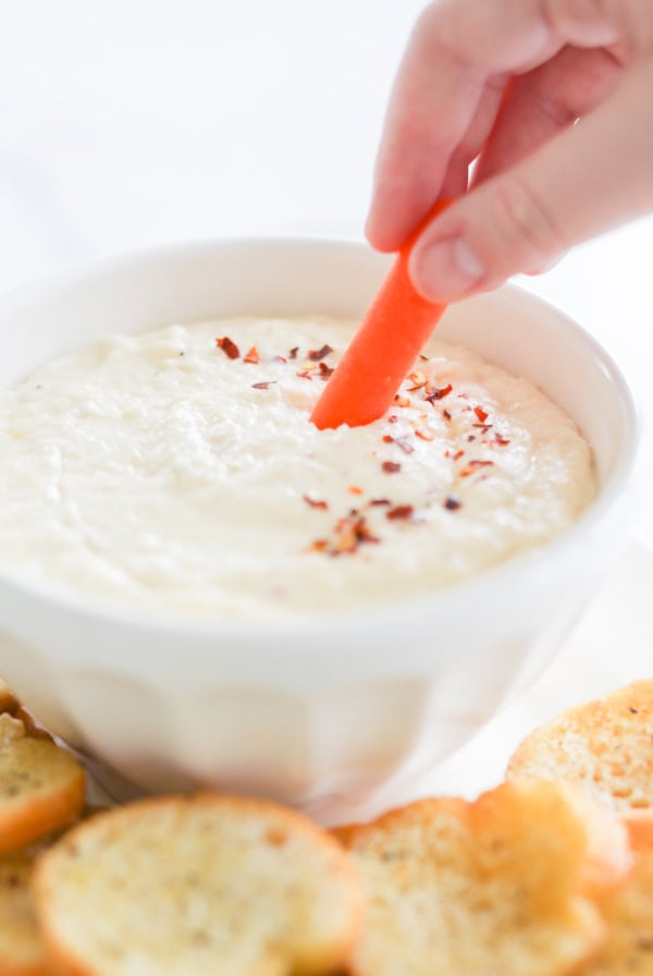 Hand dipping a carrot stick into a bowl of creamy Asiago cheese dip topped with chili flakes, surrounded by toasted bread slices.