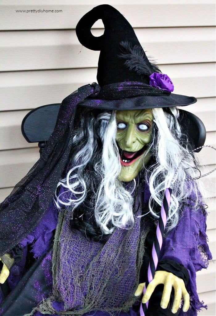 A large outdoor diy witch project sitting on a Halloween front porch. She has a green face, a very fancy witches hat, a thick mane of black and white hair and a very pretty witches hat to make her pretty and striking.