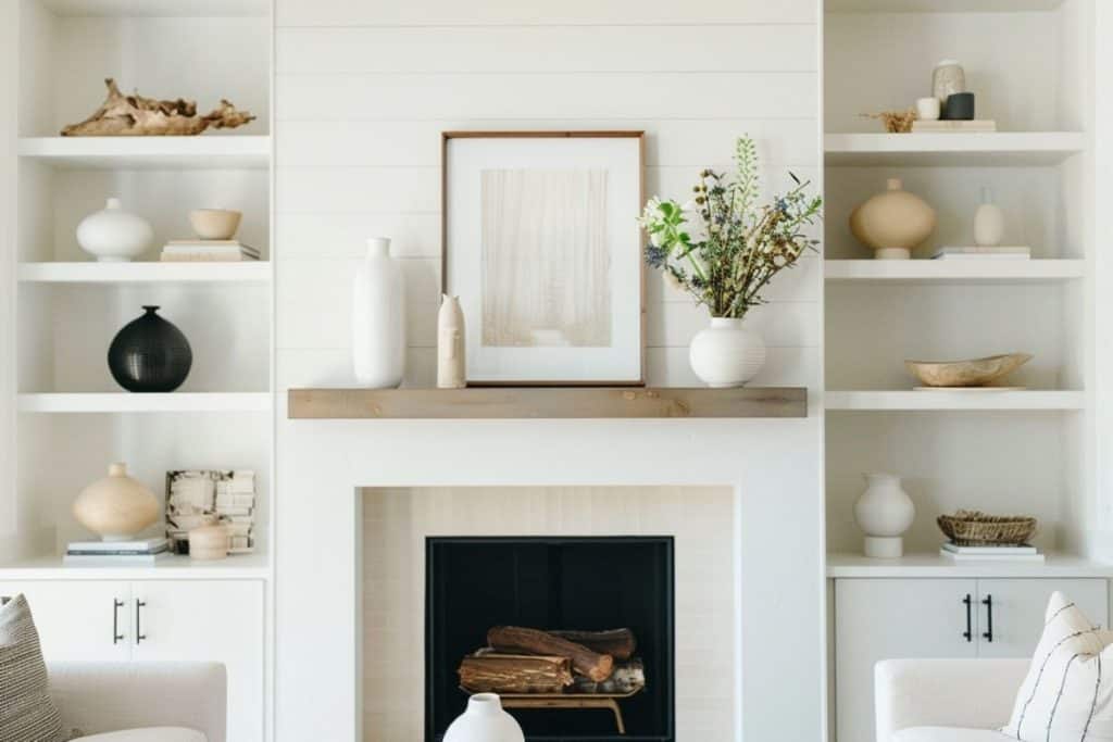 A minimalist living room with a white fireplace and built-in shelves holding neutral-toned pottery and decor. A framed art piece and a vase with flowers sit on the mantel.