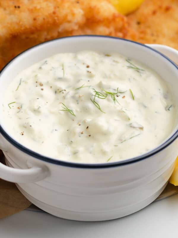 homemade tartar sauce in a white handled bowl with blue rim on a platter of fried fish with lemon wedges