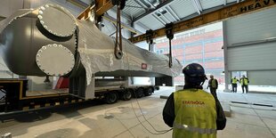 Ein Bauarbeiter mit Warnweste steht vor einem Bauteil der Flusswärmepumpe, das auf einem LKW in einer Lagerhalle liegt.