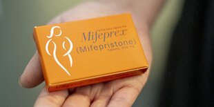 Eine orangene Packung Mifeprex. Darauf auch die Silhouette einer Person. Die Packung wird gehalten von einer flaschen Hand.