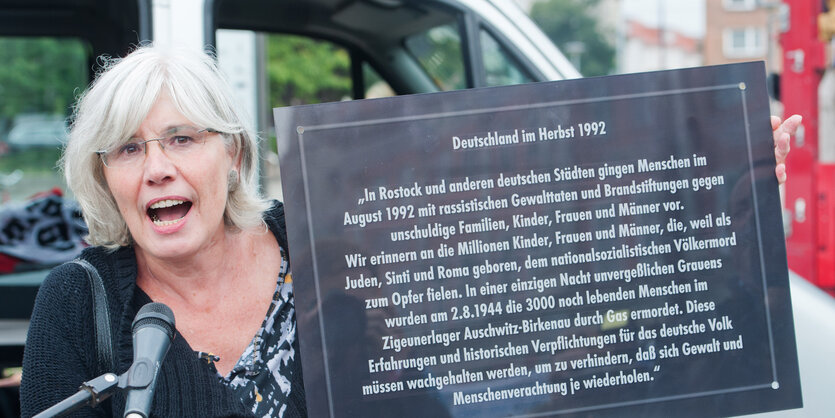 Cornelia Kerth hält vor dem Rathaus von Rostock eine Gedenktafel in der Hand und spricht in ein Mikro.