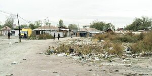 Unbefestigte Wege, an denen sich Plastikmüll zwischen verdorrter Vegetation gesammelt hat, im Hintergrund stehen Einfamilienhäuser mit roten Dachziegeln