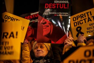 Bild von einer Demo. Es ist dunkel. Die Plakate und Gesichter sind in geld, rot, orange ausgeleuchtet oder gestaltet. Das Gesicht einer alten Frau ist näher zu sehen. Sie trägt ein Plakat.