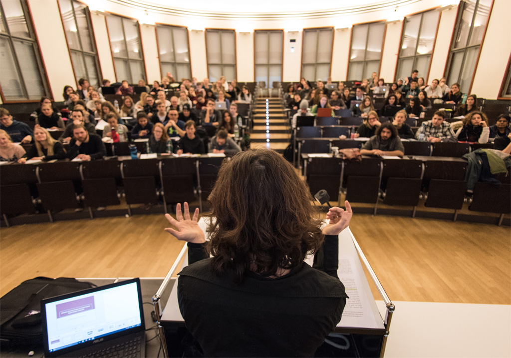 Eine Professorin steht mit dem Rücken zur Kamera und spricht mit ihren Studierenden, die im Hörsaal sitzen.