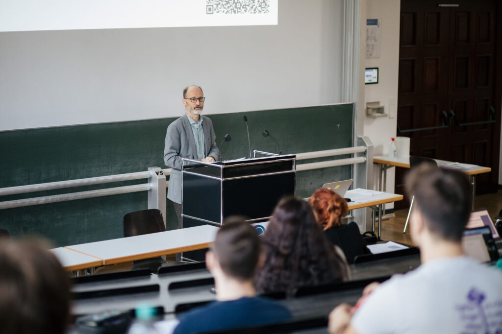 Ein Dozent steht an einem Rednerpult und hält eine Vorlesung in einem gefüllten Hörsaal der Universität Freiburg.