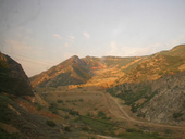 View through a dirty window, showing power lines in the foreground and a scarred mountain with the scar leading to an earthen dam.