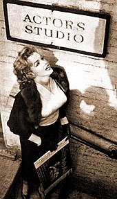 Monroe, que lleva falda, blusa y chaqueta, de pie debajo de un cartel del Actors Studio mirando hacia él.