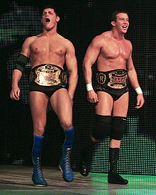 Two dark-haired men are both wearing black wrestling tights. One wears knee-high blue wrestling boots with blue knee-pads and a black elbow pad on his right arm, and the other wears black knee-high wrestling boots with black knee-pads, and white tape around his wrists. The two are wearing professional wrestling championship belts around their waists.