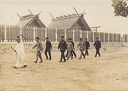 photo d'un groupe de 9 hommes en train de marcher devant le mur d'un sanctuaire. Tous portent un uniforme militaire, sauf celui en tête qui porte une tenue de prêtre shinto. Des toitures de bâtiments sont visibles derrière ce mur.