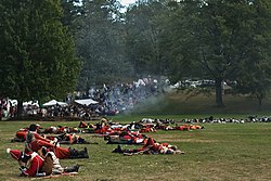 An open grassy area is strewn with bodies, most of them in red and white uniforms, although some with blue coats are visible farther back. In the distance there are white tents and a crowd of people, and a small thin cloud of smoke obscures the view a little.