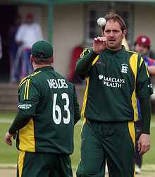 A colour photo of two cricketers, one with their back to the camera, and one looking toward the camera while catching a cricket ball.