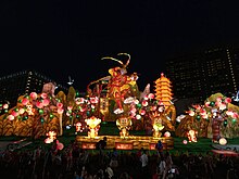 Night time image with brightly coloured lanterns and illuminated figures depicting the Monkey King in a characteristic pose surrounded by peach trees. Three smaller monkey figures depicting "see no evil", "hear no evil", "say no evil" in the foreground. Behind the decorations are high-rise buildings of downtown Singapore.