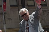 Man wearing gray jacket, tie, and sunglasses with his left arm raised