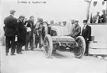 Photographie en noir et blanc d'une ancienne voiture de course, avec de nombreuses personnes à côté.