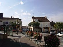 Vue d’une placette bordée de maisons de villes et fleurie.