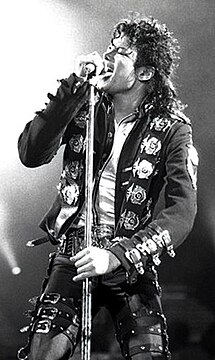 An African-American man with shoulder length black curly hair wearing a black jacket and pants adorned with buckles singing into a microphone with his eyes closed. He is holding the microphone stand with both hands. Bright stage lights are visible in the background.