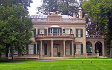 A two-story ornate brown stone house with a flat roof, dark green wooden window shutters and a semicircular front entrance porch with columns, seen from slightly to its left with some trees in front