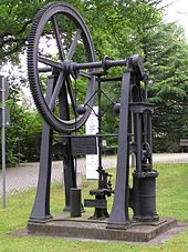 Schwarz lackierte Maschine aus Metall die auf einem Ausstellungsgelände im Freien steht. Ein Zahnrad mit mehr als einem Meter Durchmesser ist über eine Kurbelwelle mit einem Druckzylinder (etwa 40 cm hoch und 20 cm breit) verbunden. Über kleine Zahnräder ist ein Drehzahlbegrenzer verbunden. Die Maschine hat kein Gehäuse und ist etwa 2 Meter hoch.