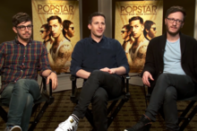 Three men (The Lonely Island) sitting on chairs in front of posters for their movie, "Popstar: Never Stop Never Popping".