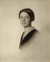 Black and white photograph of the bust of a young white woman before a white background with dark hair parted in the middle and tied up in the back, a serious countenance with eyes on the viewer, and a dark jacket over a white blouse.