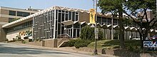 A large TV station building. The building has many windows and a large awning runs parallel to one side. A small sign out front and a large sign on the side of the building both say KWQC TV 6