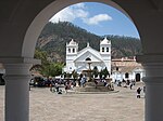 View through an arch onto a paved square with a fountain in the center and a white church on the far side