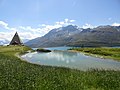 La Piramide-Chiesa della Piana delle Fontanette con davanti uno stagno. Sullo sfondo il Massiccio del Monte Giusalet e il lago. A destra, sotto al Monte Malamot (sulla destra del Giusalet), sull'altura del Crin si può notare il Forte Varisello