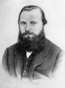 A man in his late 20s or early 30s with dark hair and a bushy beard, wearing a dark coat, dress shirt and tie.