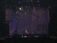 A stage with a big screen display showing a man with a beard. Sitting in a piano, a man with long curly hair wears a white shirt, with the sleeves roll back, and a black vest, plays the piano. To the man's left, a man can be seen standing playing an electric piano.