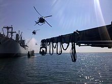 A battleship fires its guns while surrounded by helicopters and a crane.