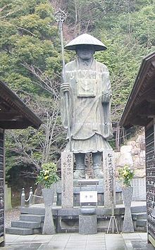 freistehende männliche Statue in Mönchsrobe mit großem konischen Hut, einem Stab in der rechten Hand und leicht erhobener linken Hand umgeben von Pflanzen und traditionellen japanischen Gebäuden