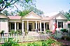 A one-story white house with green shutters, surrounded by a white picket fence, with an American flag in the front yard.