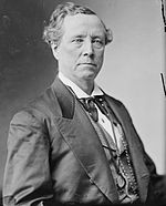 A man with wavy, receding hair, wearing a black suit with a white shirt, seated