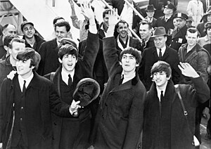 Black-and-white picture of four young men outdoors in front of a staircase, surrounded by a large assembled crowd. All four are waving to the crowd.