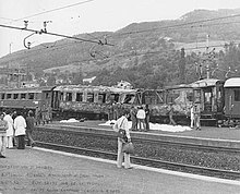 Image en noir et blanc de voitures de train défoncées et quelques passagers sur le quai.