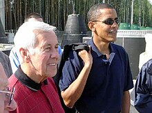 Gray-haired man and Obama stand, wearing casual polo shirts. Obama wears sunglasses and holds something slung over his right sholder.