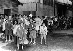 photo d'une famille regroupée le long d'un bâtiment. Ils ont des valises et des sacs pleins les mains et sont surchargés, même les enfants.