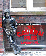 A statue depicting a young Lennon outside a brick building. Next to the statue are three windows, with two side-by-side above the lower, which bears signage advertising The Cavern Pub.