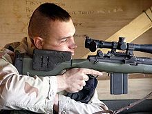 color photo of a Marine peering through the optics of a large rifle